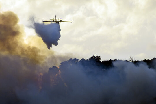 Big Fire In The Mountains With High Flames And A Long Tongue Of Fire Between Big Clouds Of Smoke And A Firefighting Plane Dumps Water On The Fire To Try To Tame It