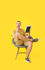 Young man reading book while sitting in armchair on color background