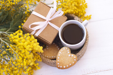 bouquet of mimosa, cup of coffee, cookies, gift box on a light background
