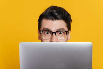 Amazed excited caucasian young adult guy with glasses peeking out from behind laptop, looking...