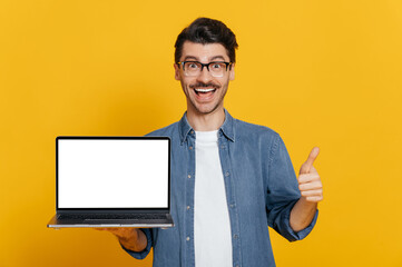 Amazed excited cheerful caucasian unshaven guy in glasses and denim shirt holds open laptop with...