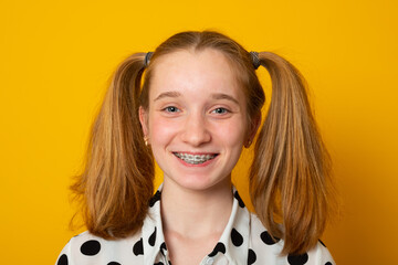 Cheerful girl with a two-ponytail hairstyle smiles with braces on a yellow background. Teenager girl with a cheerful hairstyle from two ponytails.