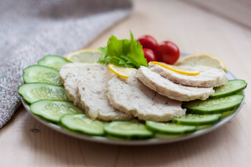 Cut the meat of a baked chicken into slices. Slices of baked chicken meat are laid out on a plate along with cucumber and cherry tomatoes.