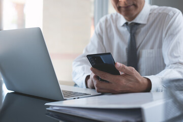 Businessman using mobile smart phone working on laptop computer with business document on table in modern office