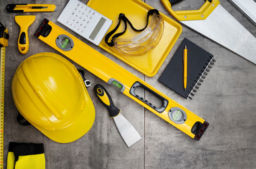 Contractor theme. Tool kit of the contractor: yellow hardhat, libella, hand saw. Plans and notebook on the gray tiles background.
