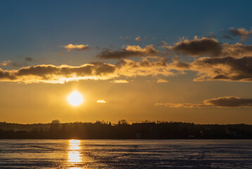 Naklejka premium Winter landscape with frozen lake at sunrise or sunset. Lake glistening ice reflect a sun.Forest in the background.
