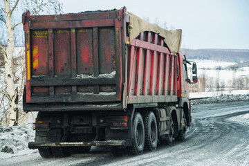 A dirty red truck carrying coal moves along a winter technological road during the day