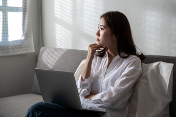 A doubt young Asian woman thinking for the future and confuses of online working on a laptop computer at home sitting on a sofa. Business home office during Coronavirus or Covid-19 quarantine concept