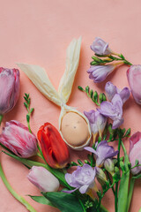DIY Easter egg with cute face and rabbit ears framed by bouquet of colorful spring flowers: tulips and freesias, on pink background. Festive springtime flat lay. Greeting card mockup. 