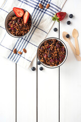 Home-made baked granola in two bowls, with berries, almonds, napkin and eco-friendly spoons. White rustic background, copy space. 