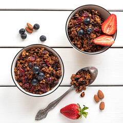 Home-made baked granola in two bowls, with berries, almonds, napkin and a spoon. White rustic background.