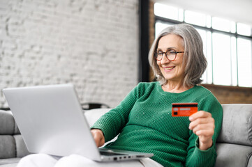 Pretty senior businesswoman in glasses and green jumper sitting at the desk using laptop and credit card for online payment from home, typing number to enter internet banking account, ordering food