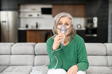 Sick senior gray-haired woman sitting on the couch in a living room, wearing an oxygen mask to...