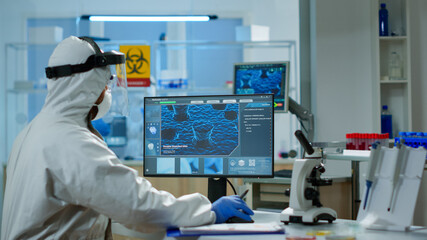 Fototapeta na wymiar Scientist in protection suit typing on pc working in medical laboratory, writing on clipboard, developing inovative drugs against new virus. Doctor using high tech, chemistry tools for researching