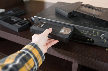 A man's hand in a checkered yellow shirt inserts an old tape cassette into a video player