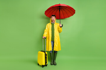 Full size photo of young man amazed hold umbrella wear raincoat baggage trip tourism flight isolated over green color background