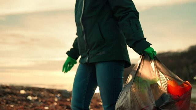 Cleaning the ocean shore. A volunteer wearing rubber gloves stands with a plastic bag in his hands and picks up a bottle from the ground. The concept of environmental protection