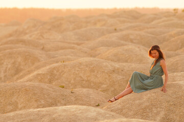 Young beautiful woman with long hair in blue dress sitting on sand at sunrise in desert. Hand touching sand