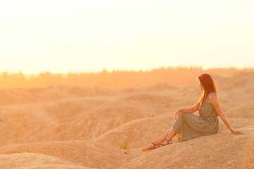 Young beautiful woman with long hair in blue dress sitting on sand at sunrise in sandy desert. Looking thoughtful down