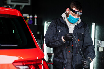 Naklejka premium Car detailing concept. Man in face mask with orbital polisher in repair shop polishing orange suv car.