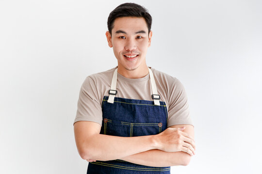 Close Up Portrait Head Shot Of Asian Short Black Hair Young Successful Handsome Male Barista Stand Cross Arms Smiling Wearing Brown Shirt And Dark Blue Jeans Bib Apron In Front White Wall Background