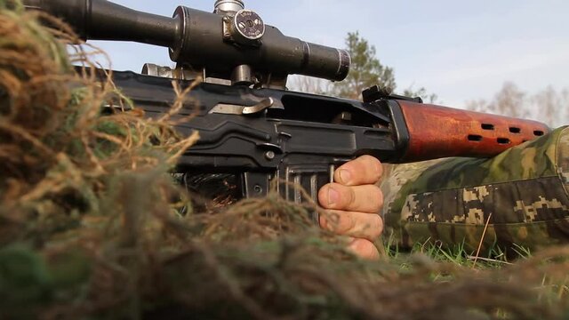 A sniper in a camouflaged sniper suit while lying down is aiming at a sniper rifle with a telescopic sight for a long time and shoots the last cartridge in the clip, close-up, Full HD, no sound.