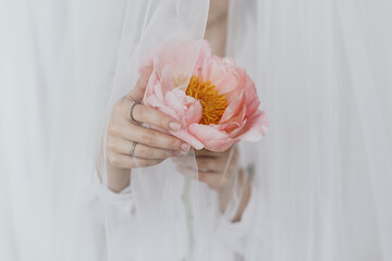 Sensual beautiful woman behind soft veil with pink peony in hands. Tender aesthetic bridal morning