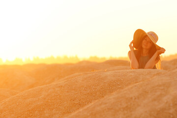 Portrait of young beautiful smiling woman with long hair with straw hat on head lying on sand at sunrise in sandy desert