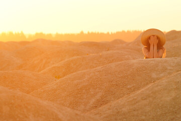 Young beautiful woman with long hair with straw hat on head lying on sand, closing face with hands. Sunrise in desert