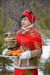 Portrait of an elegant woman in a traditional Russian winter costume holding a tray with a samovar and pies on the background of the forest