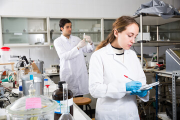 Young female scientist making research in chemical laboratory and taking notes in test chart..