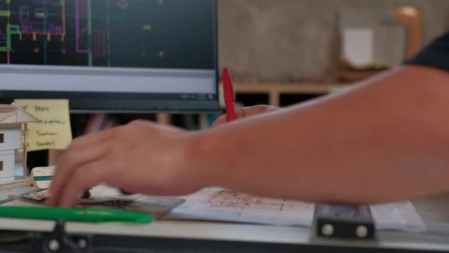 Close-up video: Architect's hand working on design work project with concentration. Using ruler & red pen to draw construction plans on drawing board in studio. And there is a computer to help work.