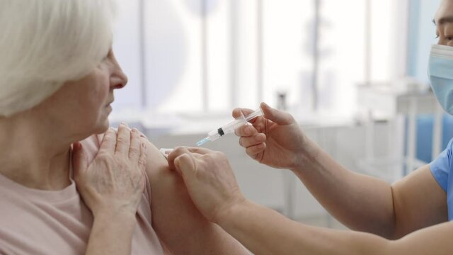 Crop view of male medical worker in protective mask disinfecting skin before giving vaccine shot in arm of retiree female person. Concept of vaccination, infectious disease prevention.