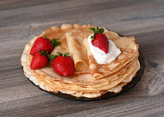 On the wooden table is a plate of pancakes decorated with sour cream and strawberries. Image with selective focus.