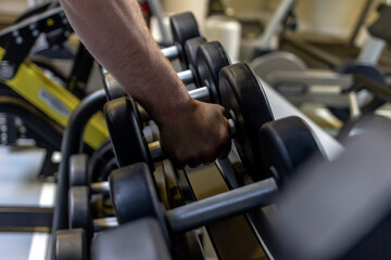 Fototapeta na wymiar Closeup view of young man grabbing a dumbbell from a dumbbell rack in the fitness center. Muscular build sportsman taking weights from a rack in a gym. Shot of a man doing weight training at the gym.