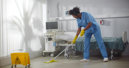 Afro-american male janitor in headphones washing floor in hospital ward