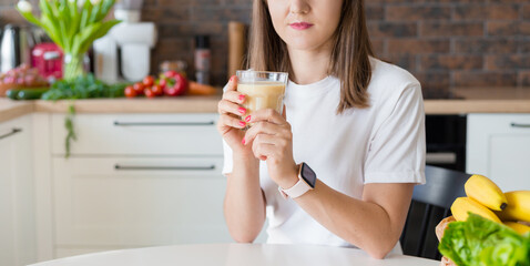 Happy brunette woman sitting with homemade glass smoothie and healthy fruits at home kitchen. Vegan meal and detox concept. Girl with white t-shirt drinking fresh cocktail