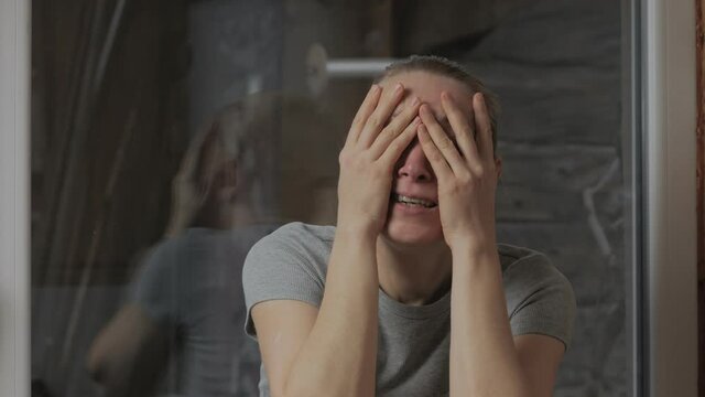 Portrait of a woman who cries sitting in the house by the window, covering her face with her hands