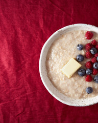 Part of oatmeal porridge with blueberry, bilberry. Close up breakfast in white bowl with fresh berries on dark red cranberry linen tablecloth. Bright color of textile. Top view, copy space, vertical 