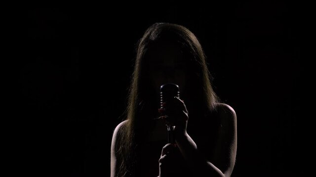 Silhouette of young woman singing into retro microphone. Female singer with long hair dances to the beat of melody and touches microphone with her hands on black studio background. Close up.
