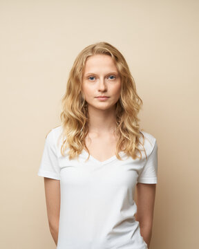 Waist up portrait of beautiful young serious clever blonde woman without makeup on beige wall. Pretty female with curly hair in white t shirt