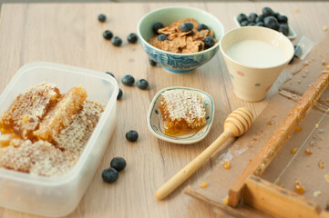 Simple healthy breakfast with cereals and blueberries in the bowl, honey in honeycomb with the spoon, cup of milk on the wooden table, selective focus