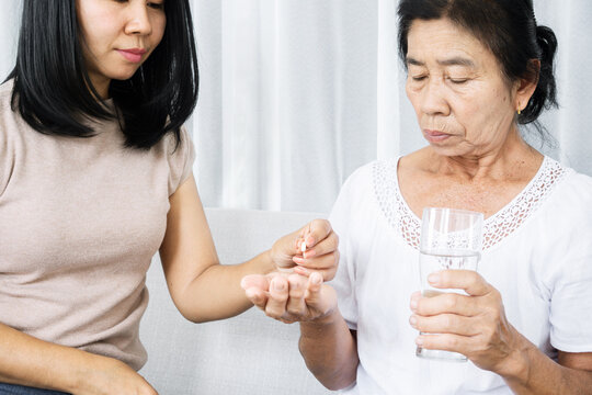 Asian Daughter Taking Care Of Old Mother Hand Giving Pills And Glass Of Water Helping Her Mom Taking Medicine On Time