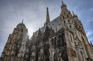 Fototapeta premium Cinematic view of St. Stephen's Cathedral in downtown of Vienna, Austria. Rays of sun between towers and tops of church. Bright sunny sky. Medieval Roman Catholic place of worship. Scenic panoramic.