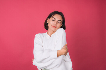 Fototapeta premium Beautiful woman in casual white sweater on pink red background love concept, holding hands crossed hugging herself isolated copy space closed eyes