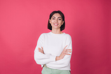 Beautiful woman in casual white sweater on pink red background with crossed hands smile look to...
