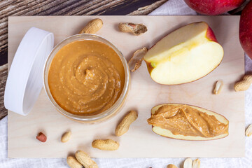 Top view of fresh homemade crunchy peanut butter with nuts and apples on light wooden background in the kitchen.