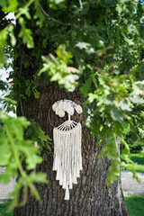 A round wicker macrame panel in the form of a tree with leaves with wooden beads hangs in the park. Natural color threads