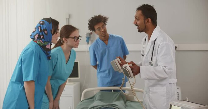 Mature indian doctor showing young interns how to use defibrillator in hospital ward