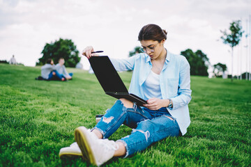 Smart female student in optical eyeglasses reading compendium preparing to school exams sitting at grass and thinking, contemplative hipster girl in spectacles thinking about education while studying